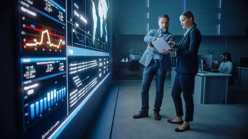 manager and computer science engineer talk while using big screen display and a laptop